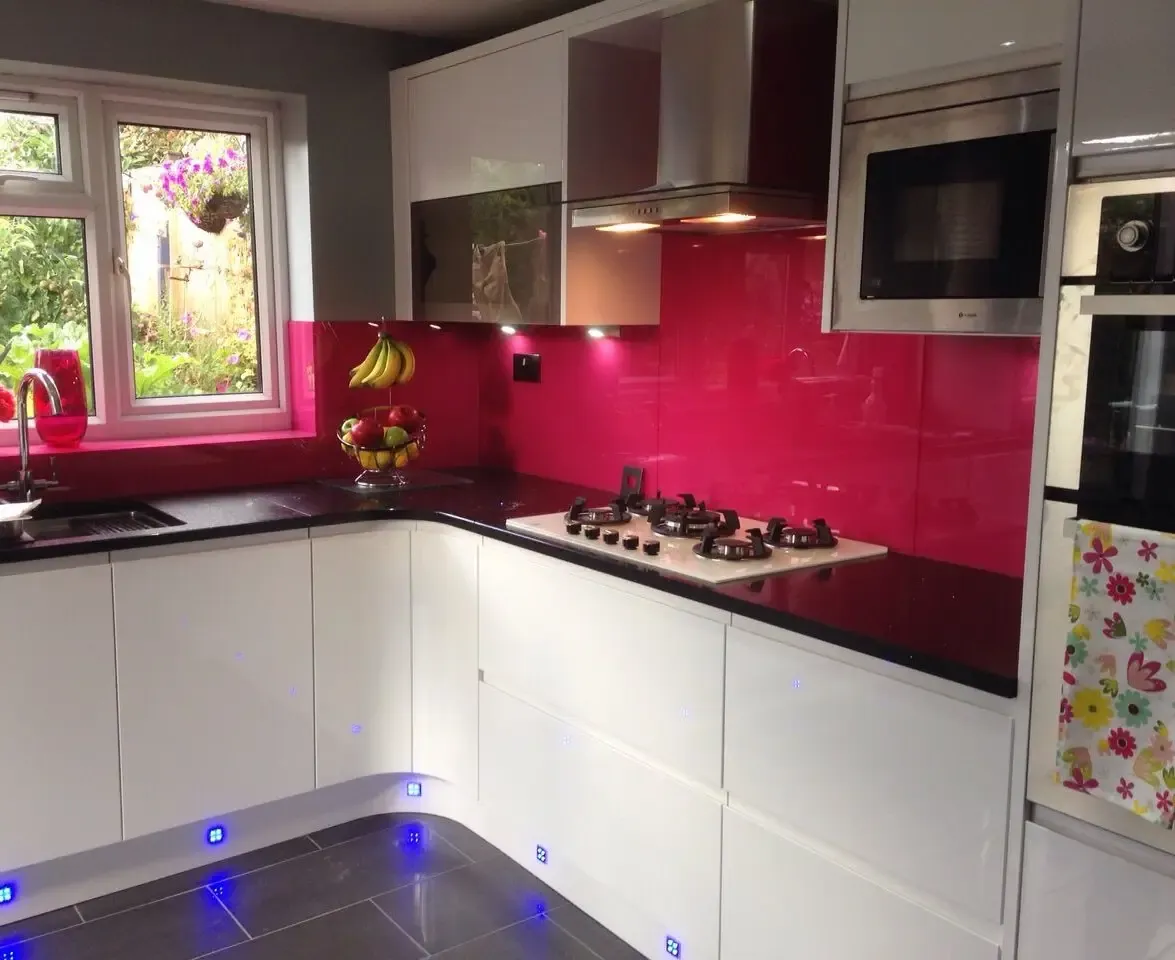 White gloss handleless kitchen with a raspberry pink glass splashback, black sparkle worktops and blue plinth lights