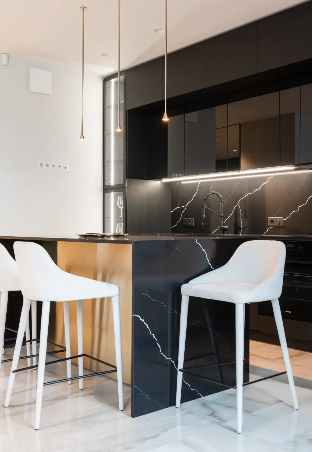 Black marble-effect waterfall island with a brass side panel, white bar stools, black gloss wall units and an LED-lit glass display cabinet